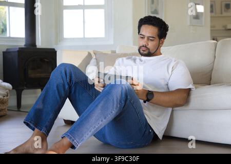 Mitte erwachsener afroamerikanischer Mann sitzt auf hellem Hartholzboden, lehnt sich gegen Sofa und verwendet ein Tablet Stockfoto