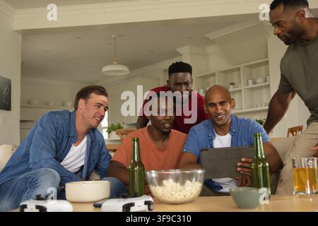 Verschiedene männliche Freunde lehnen sich vor und schauen auf ein Tablet auf dem Sofa im Wohnzimmer mit Popcorn, Controllern Stockfoto