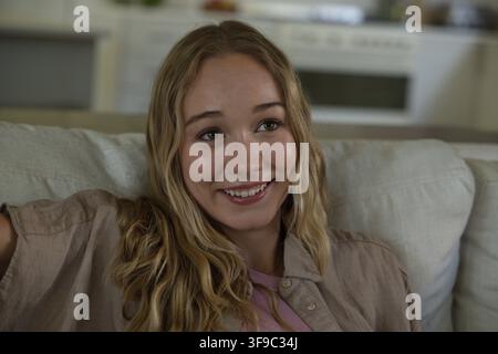 Frau in den 20ern, die auf einem hellen Sofa zu Hause sitzt und lächelt, trägt ein Hemd über einem pinken Top, Herd Stockfoto