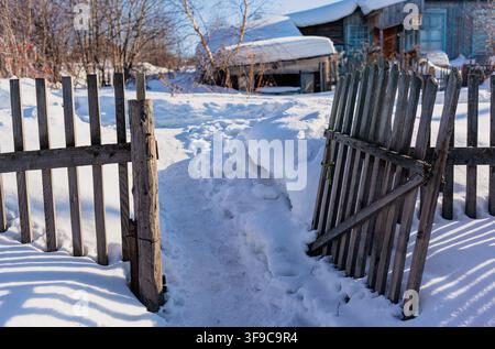 Schneebedeckter Weg durch ein offenes Holztor, der zu einem rustikalen Haus in einer Winterlandschaft mit Bäumen und Schneeverwehungen führt Stockfoto