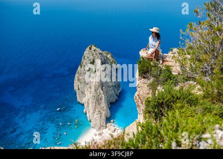 Eine Person sitzt auf einer felsigen Kante mit Blick auf das atemberaubende türkisfarbene Wasser von Myzithres, Zakynthos, Griechenland. Die natürliche Schönheit und die ruhige Atmosphäre schaffen eine perfekte Auszeit. Stockfoto