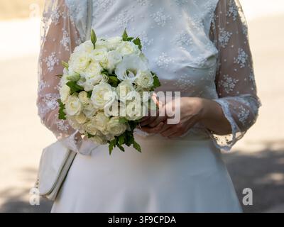 Braut hält einen weißen Rosenstrauß in einem Spitzenhochzeitskleid während einer Zeremonie im Freien Stockfoto