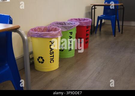 Drei Recyclingbehälter stehen an einer cremefarbenen Wand auf einem holzoptischen Boden in der Nähe von Schülerisch und -stühlen Stockfoto