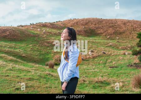 Friedliche schöne asiatische Frau steht auf grünem Hügel mit geschlossenen Augen und atmet frische Luft, umgeben von einer Schafherde, die auf den sanften Landstraßen grast Stockfoto