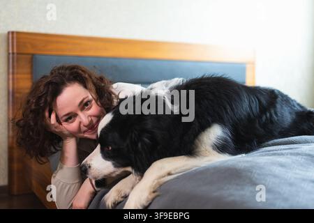 Lächelnde Frau, die mit niedlichem Hündchenhund Border Collie auf dem Bett zu Hause spielt. Besitzerin streichelt Hundefreund, der auf Sofa im Wohnzimmer sitzt. Liebes-Tier-Freundschaft-Team-Konzept Stockfoto