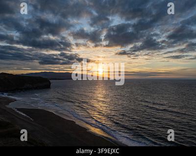 Luftaufnahme von Praia do Areal de Santa Barbara bei Sonnenuntergang mit goldenem Licht, das sich auf den Wellen des Atlantischen Ozeans spiegelt, und dramatischen Wolken über der Küste Stockfoto