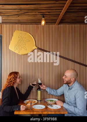 Warme Holzwände umrahmen eine gemütliche Restaurantszene. Paar prostet mit Wein an und teilt freudige Lächeln. Pizzascheibe hängt kunstvoll über ihrem Tisch. Weiche l Stockfoto