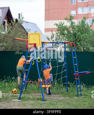 Zwei Kinder spielen auf bunten Außenspielgeräten in einer grasbewachsenen Fläche, mit einem Wohngebäude und Bäumen im Hintergrund Stockfoto