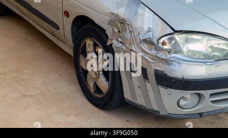 Ein silbernes Auto steht auf einem Parkplatz. Seine vordere Stoßstange ist mit Klebeband bedeckt, das eine große Delle verbirgt. Der Boden ist aus Beton und scheint draußen zu sein Stockfoto
