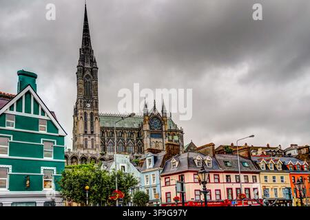 Die Türme der St Colman's Kathedrale ragen über farbenfrohe historische Häuser unter dramatischen Wolken in der Hafenstadt Cobh, County Cork. Stockfoto