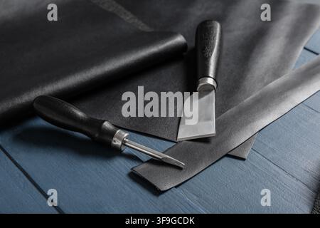 Lederhandwerkzeuge und schwarzes Ledermaterial auf einem dunkelblauen Holzgrund, handgefertigte Kunstwerkstatt Stockfoto