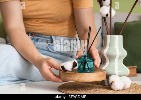 Frau arrangiert einen Schilfrohr-Diffusor und Baumwoll-Dekor auf einem Holztablett für Heimduft und Innendekoration, die blaue Glas-Diffusorflasche enthält blac Stockfoto
