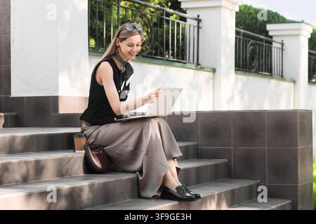 Junge Frau arbeitet remote an einem Laptop im Freien, Digital-Nomad-Lebensstil und Freelance-Karrierekonzept, eine junge Frau arbeitet remote an einem Laptop, während Stockfoto