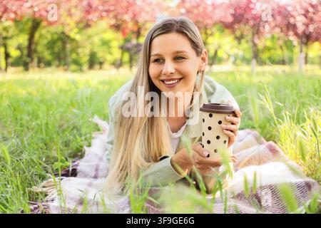 Lächelnde junge Frau entspannt sich auf einer Picknickdecke im Park, hält eine wiederverwendbare Kaffeetasse und genießt Natur und Freizeit Stockfoto