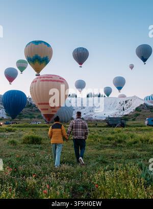 Paar schlendert Hand in Hand durch üppige Felder, während farbenfrohe Heißluftballons den Himmel über Pamukkale, Türkei, füllen. Dieser malerische Ort ist berühmt für seine Travertinterrassen. Stockfoto
