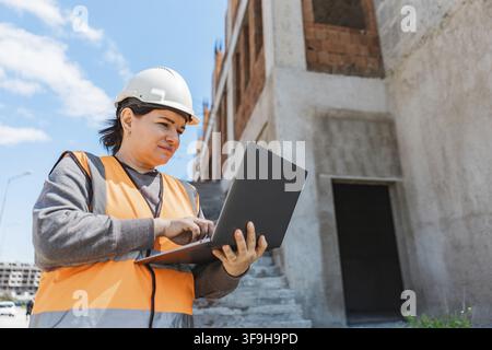 Frau mit Schutzhelm und Sicherheitsweste verwendet einen Laptop auf einer Baustelle. Das Gebäude ist im Bau mit freigelegten Ziegeln und Beton Stockfoto
