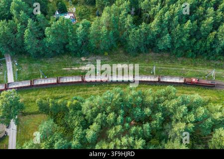 Luftaufnahme eines Güterzugs mit leeren offenen Wagen, der durch einen dichten grünen Wald in Vilnius, Bezirk Vilnius, Litauen fährt. Stockfoto