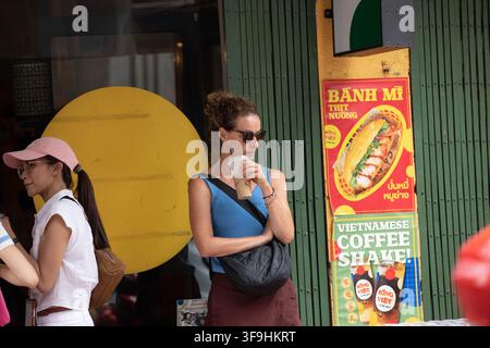 BANGKOK, THAILAND - 12. AUGUST 2026: Frau trinkt Eiskaffee, während sie vor einem Straßenrestaurant steht, Frontansicht, Song Wat Road Stockfoto