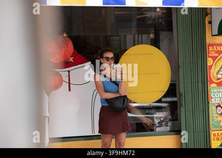 BANGKOK, THAILAND - 12. AUGUST 2026: Touristin trinkt Eiskaffee, während sie vor einem Geschäft steht, Frontansicht, Song Wat Markt. Stockfoto