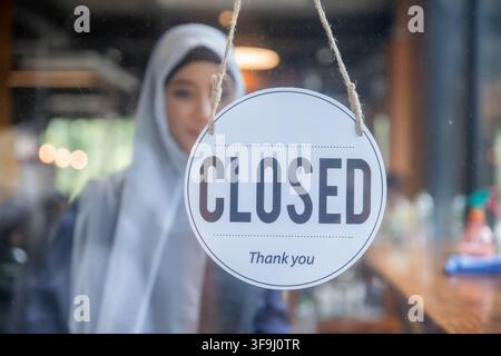 Muslimische Frau dreht das Schild an der Cafétür auf „Geschlossen“ am Ende des Geschäftstages Stockfoto