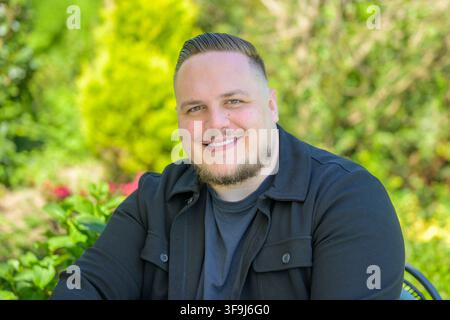 Freundlicher junger Mann, der selbstbewusst lächelt, während er im Freien in einer grünen Gartenumgebung sitzt. Natürliches Lebensstilporträt mit entspannter Atmosphäre und so Stockfoto