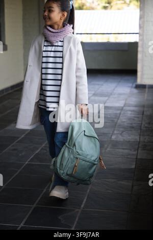 Afroamerikanischer Teenager in hellem Mantel geht durch eine überdachte Passage und trägt einen mintgrünen Rucksack Stockfoto