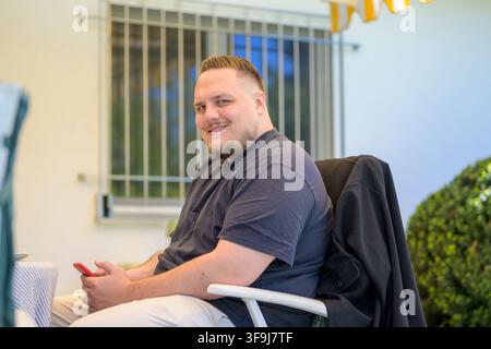 Entspannter junger Mann sitzt draußen auf einer Terrasse, während er ein Smartphone hält und natürlich in die Kamera lächelt. Lässiges Lifestyle-Porträt mit modern Stockfoto