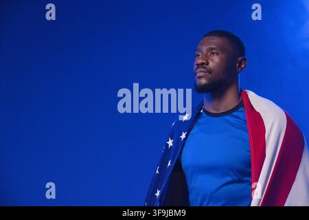 Afroamerikaner steht drapiert mit US-Flagge, blaues Sportshirt, Studionebel, Kopierraum Stockfoto