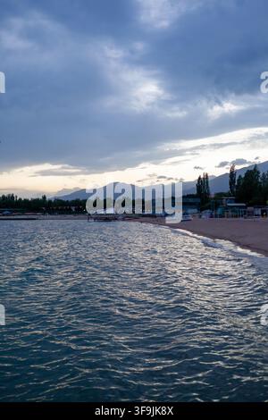 Issyk Kul, Kirgisistan – 01. Juli 2022: Ruhige Seeblickansicht mit ruhigem Wasser, sandigem Strand und fernen Bergen unter einem bewölkten Himmel in der Dämmerung Stockfoto
