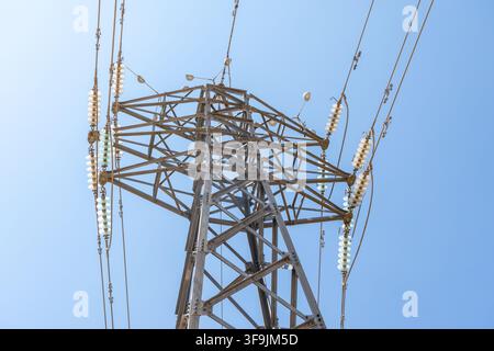 Diese detaillierte Aufnahme konzentriert sich auf die komplexe Metallstruktur eines Übertragungsmastes und hebt die Isolatoren und Drähte vor einem hellen, wolkenlosen Hintergrund hervor Stockfoto
