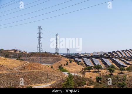 Reihen von Photovoltaikpanelen sind auf einem Hügel installiert, mit hohen Strommasten und Übertragungsleitungen, die sich vor einem klaren blauen Himmel abheben Stockfoto