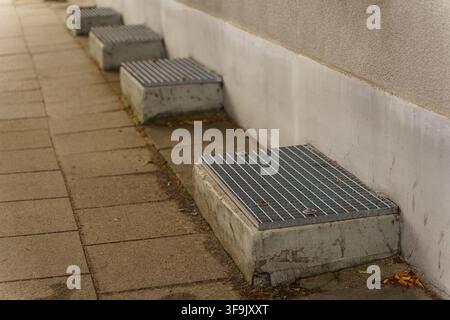 Reihe von Metallabflussgittern, die auf Betonblöcken entlang einer Gebäudemauer auf einem gefliesten Gehweg montiert sind, repräsentiert städtische Infrastruktur, Entwässerungssystem Stockfoto