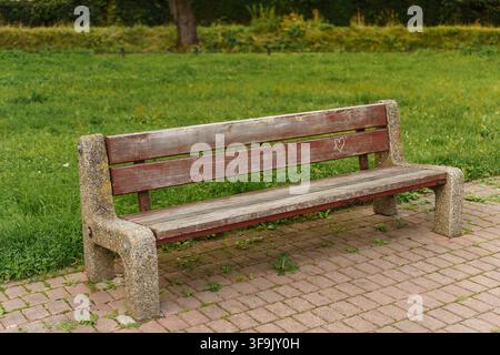 Eine gealterte, verwitterte Holzbank mit Betonstützen, die auf einem gepflasterten Steinweg liegt, vor einem Hintergrund aus grünem Gras und Laub, Entspannung im Freien, Sitzplatz im öffentlichen Garten Stockfoto