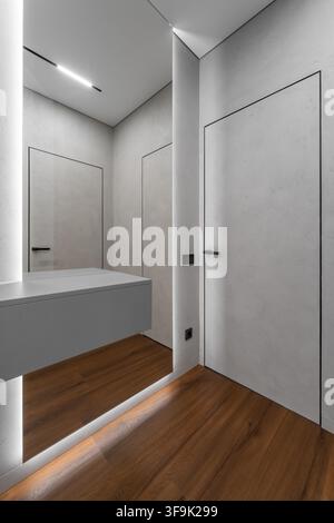 Minimalistischer moderner Apartmentflur mit verdeckten Türen, großem Wandspiegel, warmem Holzfußboden und integrierter Linearbeleuchtung Stockfoto