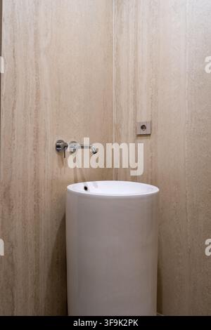 Minimalistisches modernes Badezimmer-Interieur mit einem weißen freistehenden Waschtisch, wandmontiertem Chrom-Wasserhahn und warmen beigen texturierten Steinwänden Stockfoto