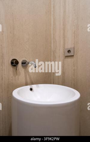 Elegantes zeitgenössisches Badezimmer mit einem skulpturalen weißen Waschtisch, minimalistischem Chrom-Wandhahn und natürlicher beiger Travertin-Wandverkleidung Stockfoto
