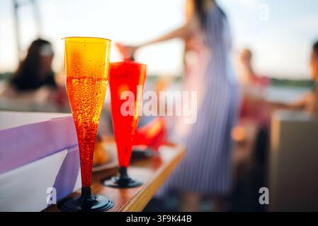 Zwei leuchtend orangefarbene Flöten, gefüllt mit einem sprudelnden Getränk, stehen auf einem Holztisch, mit verschwommenen Figuren von Leuten, die im Hintergrund gesellig sind Stockfoto