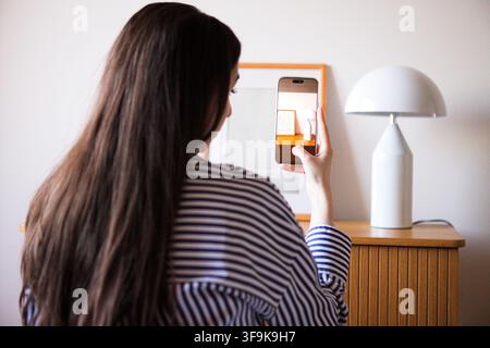 Frau mit langen Haaren nimmt ein Bild mit ihrem Smartphone auf, während sie an einem Holztisch sitzt, im Hintergrund eine moderne Lampe und Kunstwerke Stockfoto
