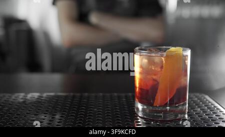 Negroni-Cocktail mit einer lebendigen Orangenhaut auf der Theke, Barkeeper im verschwommenen Monochrom-Hintergrund Stockfoto