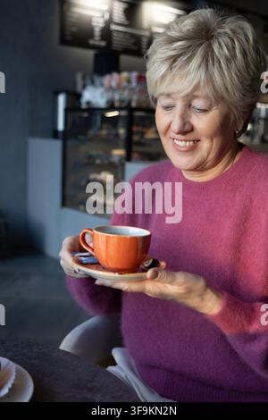 Elegante reife Frau mit zeitloser Schönheit lächelt, während sie eine Tasse heißen Kaffees in einem modernen schwedischen Café hält. Stockfoto