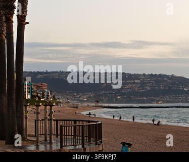 Strandszene mit Palmen und Meeresblick. Menschen schlendern oder schwimmen im ruhigen Wasser. Der Sonnenuntergang wirft ein warmes Licht über die Küstenstadt. Ideal für Reisen, Urlaub, Stockfoto