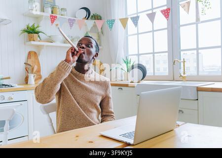 Afroamerikaner feiert seinen Geburtstag virtuell zu Hause, trägt einen Partyhut, bläst eine Partytröte und schaut auf einen Laptop in einer mit Fähnchen geschmückten Küche Stockfoto