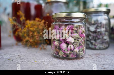 Eine Nahaufnahme eines kleinen Glases, gefüllt mit getrockneten rosa Rosenknospen für Kräutertee, mit Krüglchen voller Kräuter und rotem Öl im sanft verschwommenen Hintergrund. Stockfoto