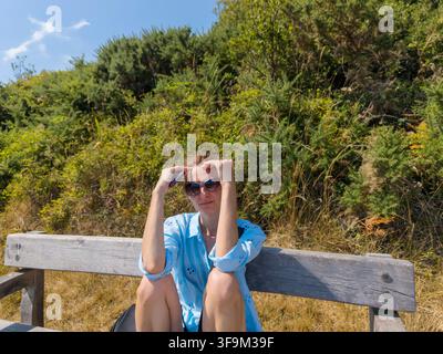 Frau sitzt auf einer Holzbank im Freien in der Natur und trägt eine Sonnenbrille und ein hellblaues Hemd Stockfoto