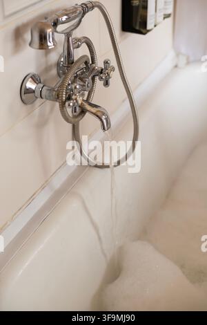 Wasser strömt aus einem vintage Chrom-Wasserhahn in eine weiße Badewanne, die mit Seifenblasen gefüllt ist, in einem hellen Badezimmerinterieur. Stockfoto