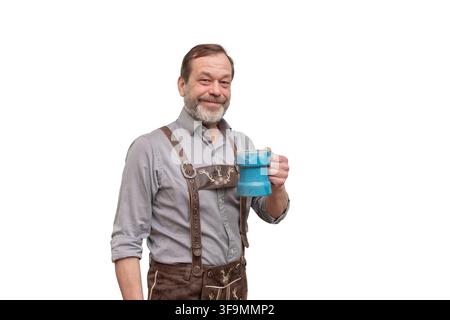 Ein lächelnder älterer Mann in traditioneller bayerischer Tracht hält einen blauen Bierkrug vor weißem Hintergrund. Stockfoto
