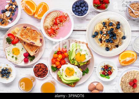 Gesunde Frühstücks- oder Brunch-Tischszene auf einem weißen Holz-Hintergrund. Blick von oben. Avocado-Toast, Smoothie-Schalen, Haferflocken, Joghurt und verschiedene Nährstoffe Stockfoto