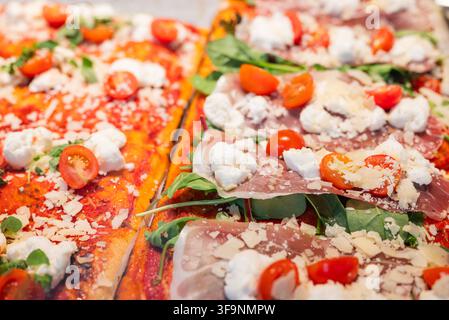 Verschiedene rechteckige Pizzastücke mit Prosciutto, Tomaten, Blattgemüse und Käse belegt, frisch an einem Café- oder Bäckerregal präsentiert. Stockfoto