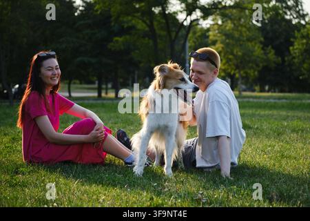 Junges Paar entspannt sich auf dem Gras mit ihrem Mischlingshund im Park zur goldenen Stunde. Haustiere, Liebe und Lifestyle im Freien Konzept. Stockfoto