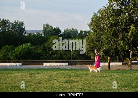 Paar genießt einen Sommer-Spaziergang mit seinem Hund im Park, umgeben von grünen Bäumen und Natur. Seitenansicht-Porträt einer glücklichen jungen Familie. Stockfoto
